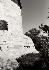 Side view of Cathcart Tower, showing the massively constructed, smooth, steep exterior walls built of tightly fitting limestone blocks and two of the four evenly spaced caponiers around the base, 1992.; Parks Canada Agency / Agence Parcs Canada, 1992.