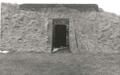View of the west Powder Magazine, showing the protected entrance flanked by brick sidewalls that slope down to ground level, 1989.; Parks Canada Agency / Agence Parcs Canada, 1989.