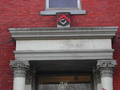 This photograph shows the entablature over the entrance, the corinthian capitals and an insignia with an arm bearing a sword, 2005; City of Saint John