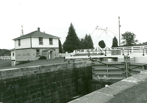 View of the Lower Brewers Defensible Lockmaster's House from a distance, showing its overall scale, design and materials, which harmonize with the lockstation surroundings, 1989.; Department of Public Works / Ministère des Travaux, 1989.