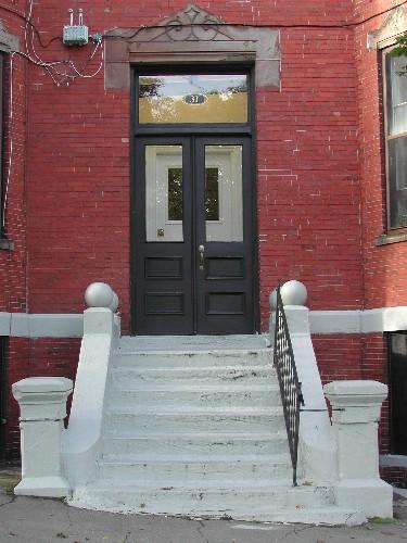 This photograph shows the entrance to the residence, and illustrates the peaked sandstone lintel, broad sandstone steps, sandstone plinth band and transom window, 2005; City of Saint John