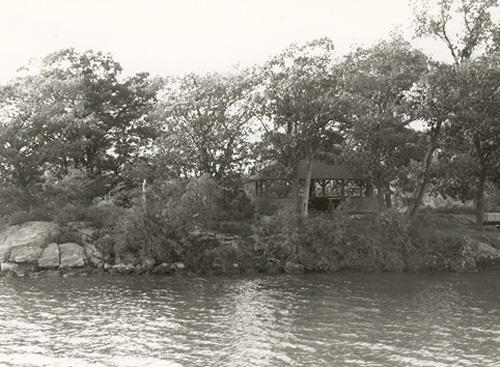 View of the Adelaide Island Picnic Shelter from the river, showing its relatively isolated island location on a heavily treed site, 1992.; Parks Canada Agency / Agence Parcs Canada / Historica Resources Ltd., 1992.