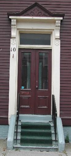This image provides a view of the wooden entablature with pediment over the entrance, brackets, pilasters, transom window and paired wooden doors with glass panels, 2005; City of Saint John