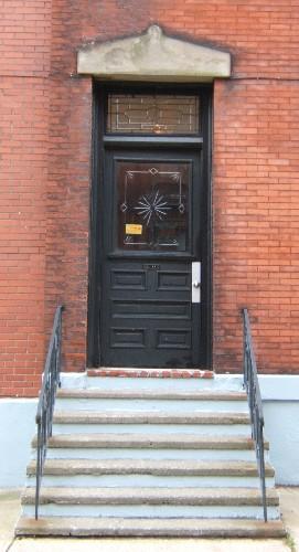 This image provides a view of the entry consisting of a pedimented, sandstone lintel above a stained glass transom window and a wood door with a glass panel, 2005; City of Saint John