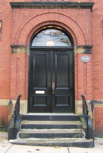 This image provides a view of the Roman arched entrance with brick pilasters crowned by Doric sandstone capitals and paired wood doors, 2005
; City of Saint John