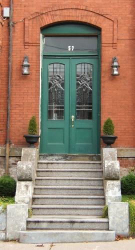 This image provides a view of the segmented arched entrance, the brick hooded lintel, the segmented arched transom window and the wooden double doors with segmented arched stain glass panels, 2005
; City of Saint John