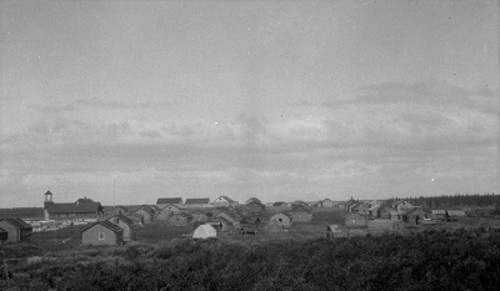 Historic photograph of Fort McPherson showing dwellings and the Anglican Church on the left of the image, standing still, ca. 1930; J. F. Moran / Indian and Northern Affairs Canada | Affaires indiennes et du Nord Canada / Library and Archives Canada | Biliothèque et Archives Canada, a102490-v8