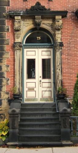 This image provides a view of the entry consisting of an elaborate sandstone entablature supported by brackets, fluted pilasters with Corinthian capitals, Roman arch transom window and paired wood doors with glass panels, 2005; City of Saint John
