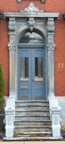 This image provides a view of the entry with the elaborate sandstone entablature supported by brackets and fluted pilasters with Corinthian capitals above a Roman arched transom window and paired wood doors with glass panels, 2005; City of Saint John