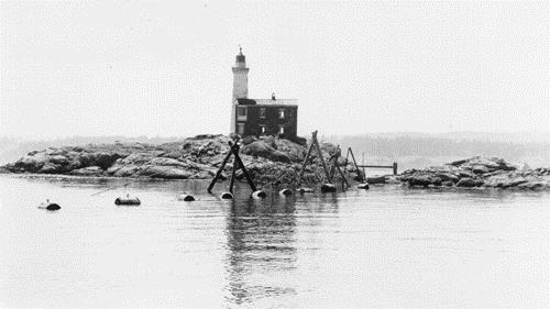 View of the back side of Fisgard lighstation during the Second World War, showing buoys holding up the submarine net. The balcony above the back door is visible on the left side of the image .; PAC, DND, RCN E-3194