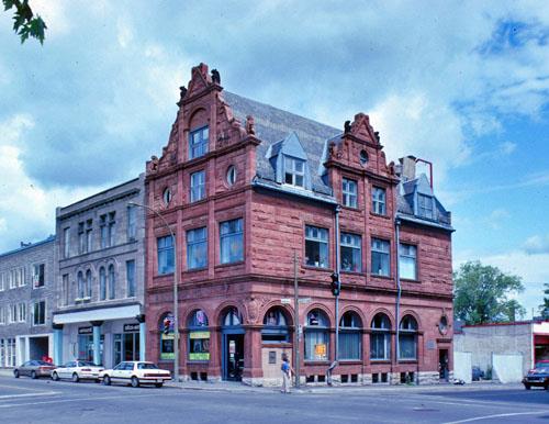 General view of the Bank of Montréal showing its use of textured, red sandstone on its two street facades.; Parks Canada Agency / Agence Parcs Canada, n.d.