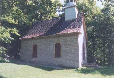 View of the main entrance to Sainte Anne Processional Chapel, showing its compact single storey massing under a steeply pitched gable roof with bellcast eaves, 1999.; Parks Canada | Parcs Canada
