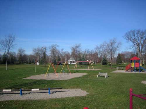 Image of Wilmot Park showing playground equipment in foreground and bandstand and Old Government House in background; City of Fredericton