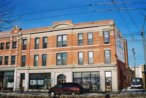 This image illustrates the front, south-facing, primary brick-clad facade of the building showing the stores at ground level with two floors of residential uses above. Note the symmetrically-designed, classically-influenced facade details.; City of Edmonton, 2004