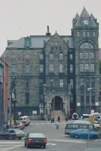 Front facade of St. John's Court House showing the three-storey, Duckworth Street facade, characterized by a central projecting bay with arched entrance, a series of blind arches, a square tower at one corner, and a massive round tower at the other.; Parks Canada/Parc Canada, n.d.