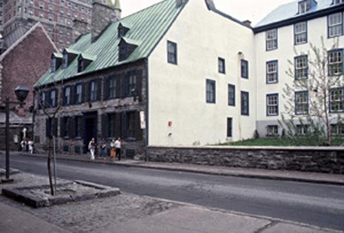 Corner view of the Maillou House showing its exterior masonry walls, the dressed stone of the door and widow frames and string course between the two storeys, and the clapboard cladding of the northeast gable, 1985.; Parks Canada Agency / Agence Parcs Canada, A. Guindon, 1985.
