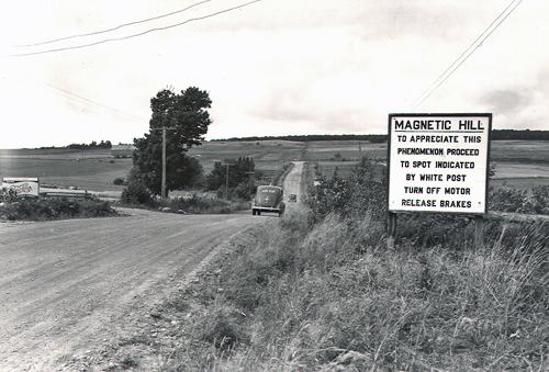 The sign in this c1940 photo reads, "MAGNETIC HILL:  TO APPRECIATE THIS PHENOMENON PROCEED TO SPOT INDICATED BY WHITE POST TURN OFF MOTOR RELEASE BRAKES"; Moncton Museum