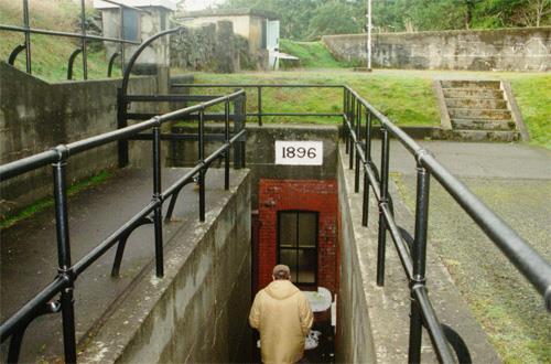 View of the main entrance to the Upper Battery Magazine showing the steps going down into the entrance well, protected by pipe railings. The window of the crew shelter is also depicted in the photograph, 1997.; Agence Parcs Canada / Parks Canada Agency, J. Mattie, 1997.