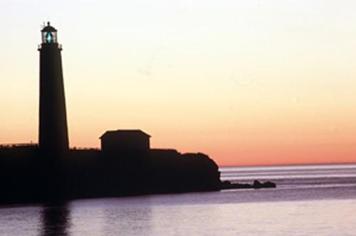 Panoramic view of the Lighttower at Cap-des-Rosiers against a setting sun backdrop putting into emphasis the elegant form and proportions of the lighttower which consists of a 112 foot tall, tapered round shaft that supports a multi-facetted lantern, 2001; Parks Canada Agency/Agence Parcs Canada, J.F. Bergeron, 2001.