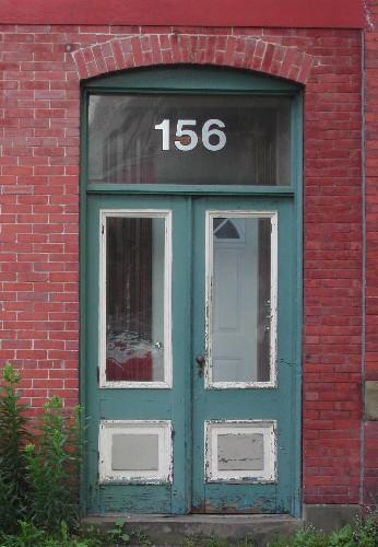 This image provides a view of the paired wood doors with glass panels below a segmented arched opening framing a rectangular transom window, 2005.; City of Saint John