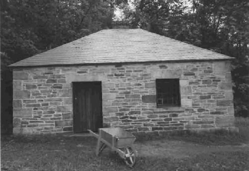 View of the main entrance to the Blacksmith's Shop, showing the simple compact, one-storey massing and the hipped roof clad in tin, 1989.; Public Works and Government Services Canada / Travaux publics et Services gouvernementaux Canada, 1989.