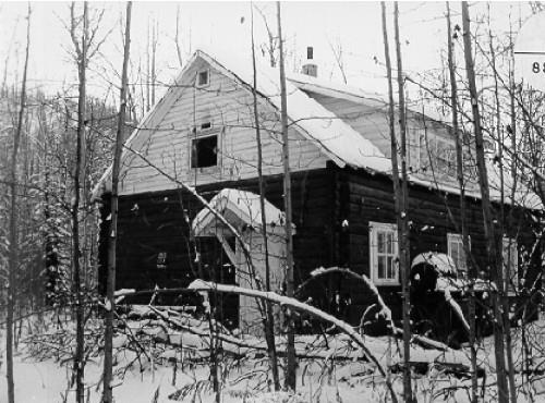 View of the main entrance to the Troberg Residence, showing its exposed saddle-notched log walls, its horizontal siding above the roof line, and its gable roof, 1988.; Agence Parcs Canada / Parks Canada Agency, 1988.